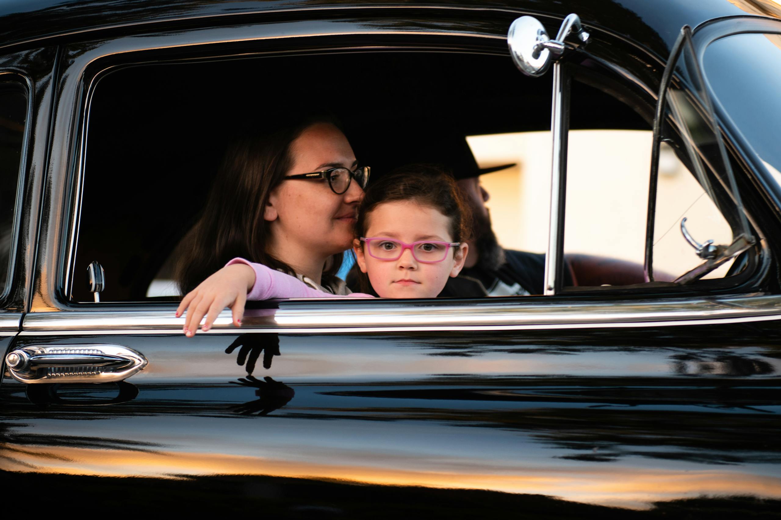 A mother and daughter enjoy a car ride in a vintage black automobile, capturing a serene family moment.